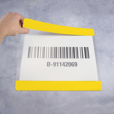 Hand hält eine transparente Beschriftungstasche mit eingelegtem Papier mit Barcode und der Kennung „B-91142069“, gelbe Randstreifen oben und unten, zur Lager- und Bodenmarkierung im industriellen Bereich.