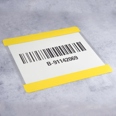Weiße Karte mit Barcode und der Kennung „B-91142069“, geschützt in einer transparenten Beschriftungstasche mit gelben Randstreifen oben und unten zur Boden- und Lagerkennzeichnung, auf hellgrauem Untergrund.