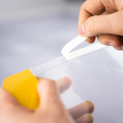 Zwei Hände bringen eine transparente Schutz- bzw. Beschriftungsfolie von einem gelben Spender auf eine glatte Oberfläche auf, wobei die selbstklebende Bodenmarkierung im industriellen Bereich angebracht wird.