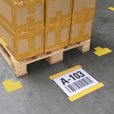 Palettenplatz mit gestapelten Kartons auf Holzpalette, umwickelt mit Folie und gelbem Band, umgeben von gelben Bodenmarkierungen. Im Vordergrund Beschriftungsschild A4 MA-WT-5118 mit gelbem Rand, Kennzeichnung „A-103“ und Barcode zur Lager- und Palettenplatzmarkierung.